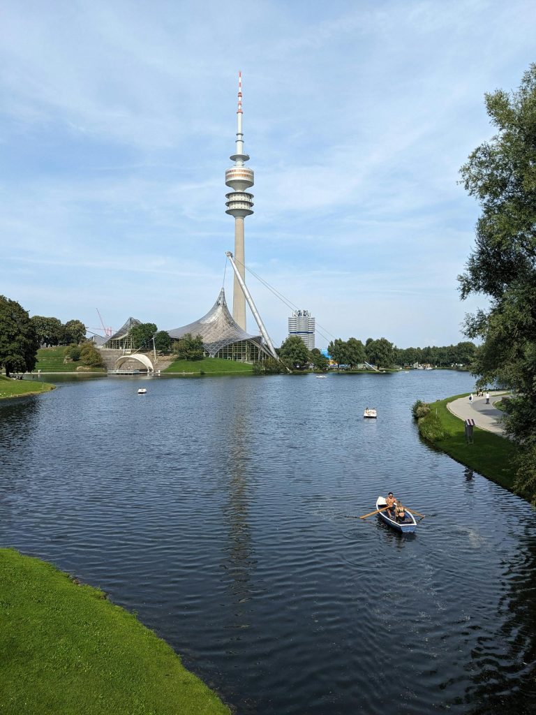 A serene summer day at Olympiapark, Munich, featuring the iconic TV tower and peaceful boating on the lake.